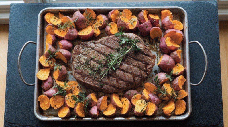Sheet pan with sliced flank steak, roasted sweet potato wedges, and charred broccolini, drizzled with green chimichurri sauce