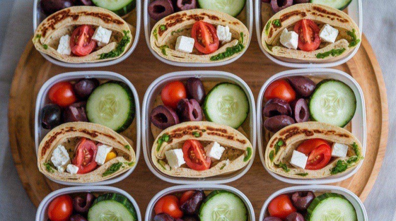Colorful Mediterranean wraps cut in half showing layers of hummus, fresh vegetables, feta cheese, and olives, arranged in clear containers