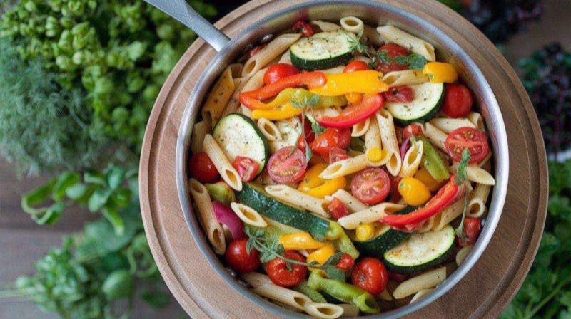 Creamy penne pasta primavera with colorful broccoli, zucchini, red peppers, and cherry tomatoes in a large pan, topped with Parmesan and basil